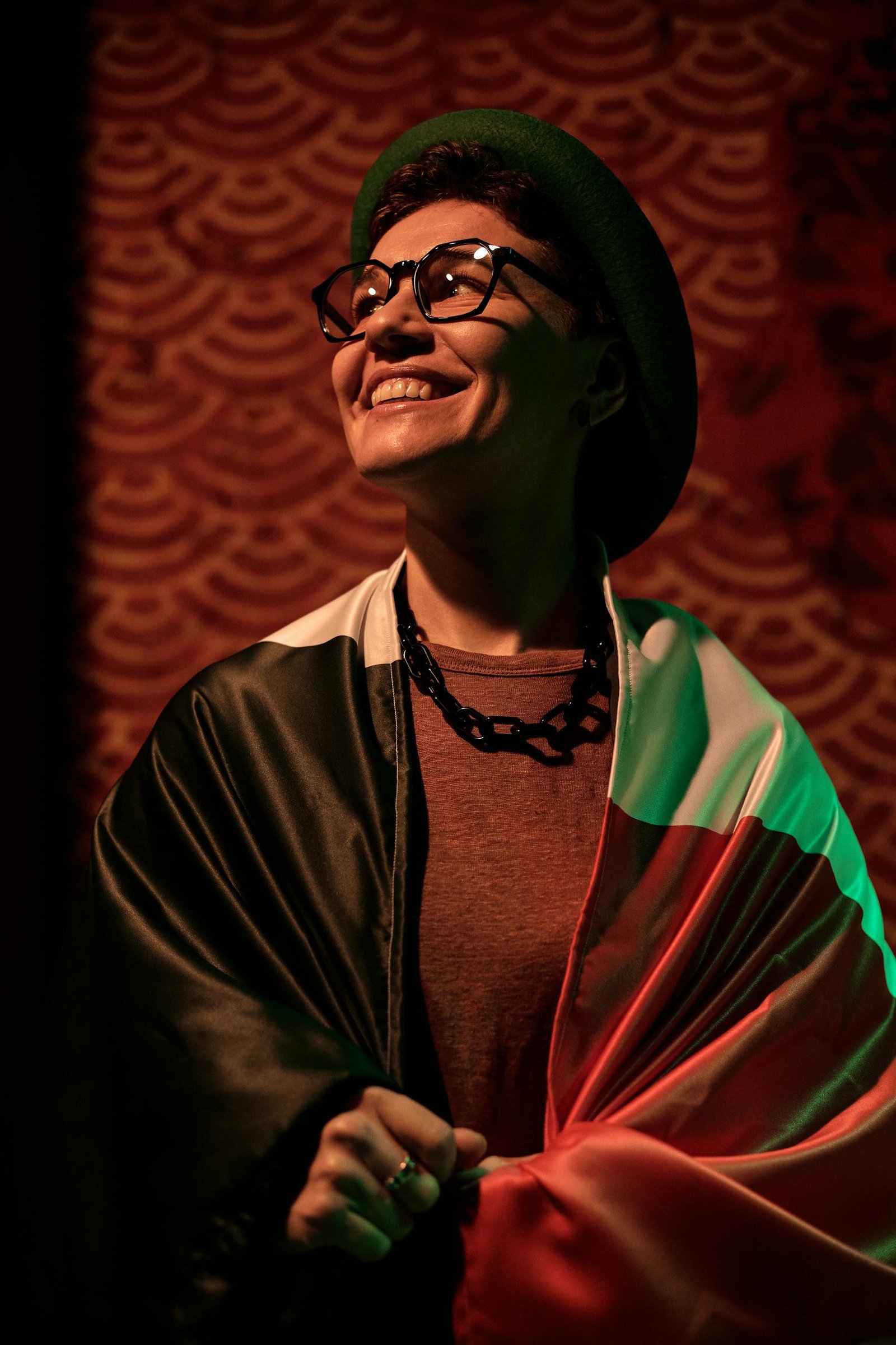 A smiling person wearing glasses and a hat, wrapped in a flag, celebrates indoors.